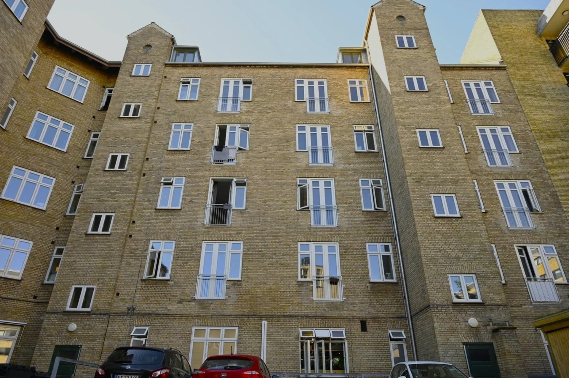 Bygningsfacade med mange vinduer, parkeringsplads og moderne boligkompleks.