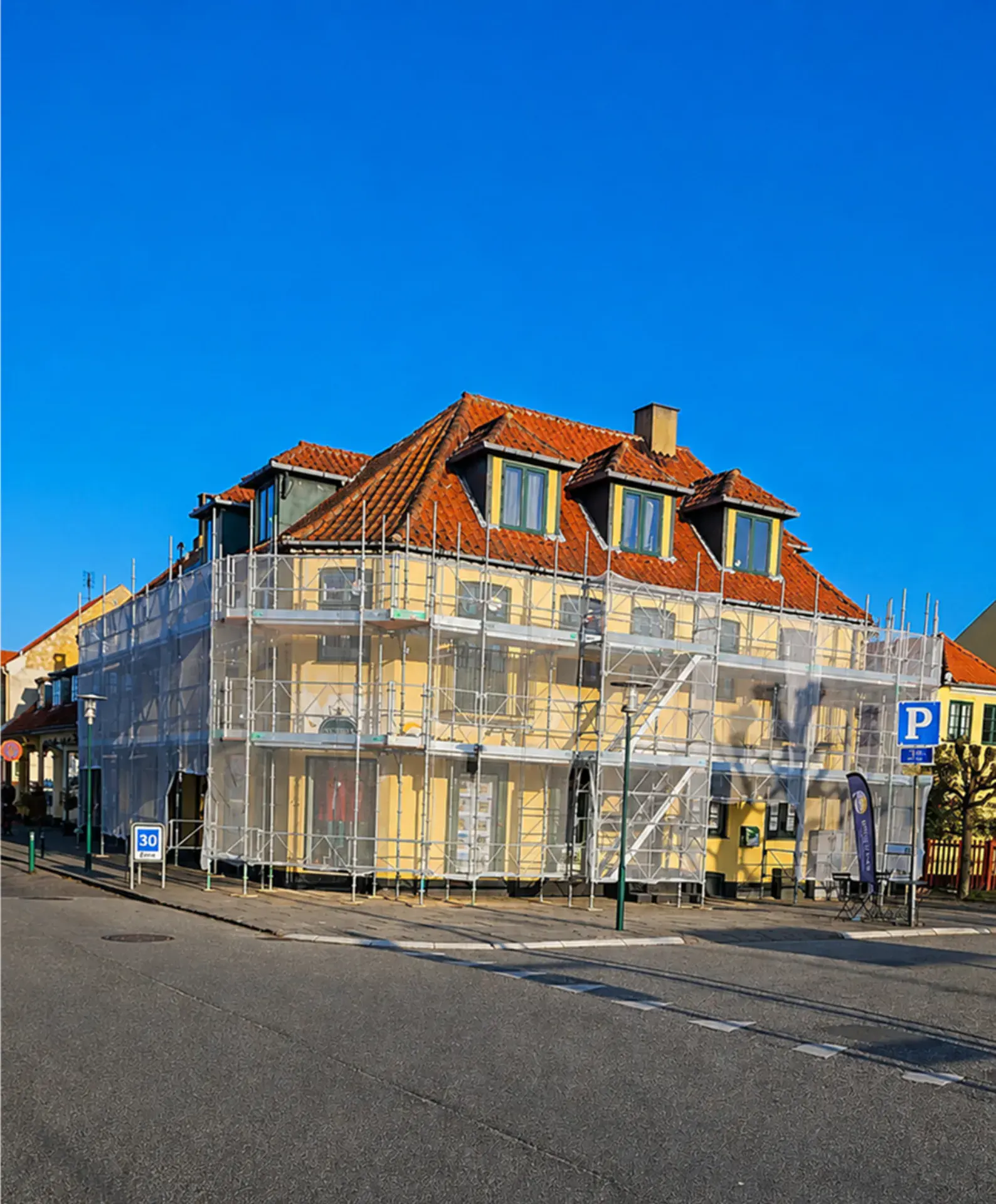Byggeri med stilladsopsætning foran et hus med rødt tag.